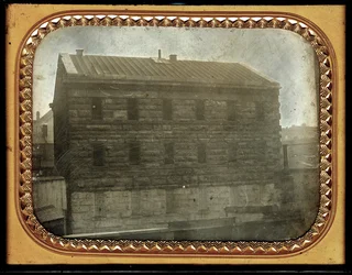 La cárcel de la ciudad en la esquina sureste de las calles Sixth y Chestnut. Pequeñas chozas de madera están cerca de la cárcel, así como cercas. Se puede ver algo de arquitectura en el fondo